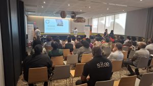 Un groupe de personnes assises dans une salle de conférence, regardant un présentateur parler à côté d'un écran de projection affichant les détails de l'événement.