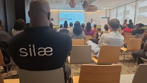 Public assis dans une salle de conférence face à un écran de présentation ; une personne au premier plan porte une chemise avec le texte 