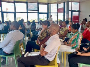 Un groupe de personnes assises sur des chaises colorées dans une pièce lumineuse, écoutant attentivement une présentation ; certaines prennent des notes, et des fenêtres bordent l'arrière-plan.