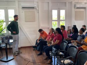 Un homme est debout et s'adresse à un groupe de personnes assises dans une pièce lumineuse au sol en bois et aux murs blancs. Un ordinateur portable et un projecteur sont placés devant lui.