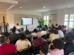 Un groupe de personnes assises dans une salle écoute une présentation. Deux présentateurs sont assis près d'un grand écran affichant une feuille de calcul.