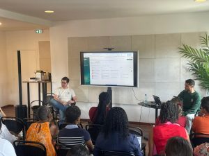 Un groupe de personnes est assis face à deux présentateurs ; l'un parle tandis que l'autre utilise un ordinateur portable. Un grand écran affiche une page web dans une salle bien éclairée.