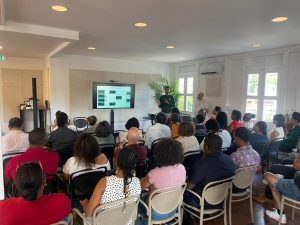 Un groupe de personnes assises dans une salle écoute un présentateur debout à côté d'un écran affichant une présentation.