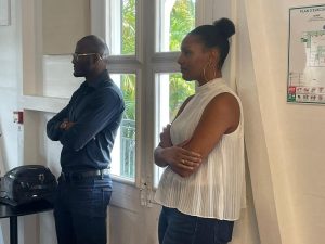 Deux personnes se tiennent à l'intérieur, les bras croisés, dans la même direction. Une fenêtre et un plan d'évacuation mural sont visibles à l'arrière-plan.