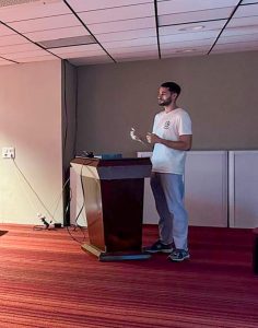 Un homme vêtu d'un tee-shirt blanc et d'un pantalon gris se tient debout et parle sur un podium dans une pièce à la moquette rouge et au plafond carrelé.