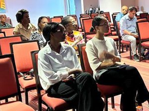 Dans une salle de conférence ou de réunion, des personnes sont assises sur des chaises rouges et écoutent attentivement. Certains prennent des notes tandis que d'autres regardent vers l'avant de la salle.