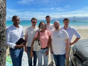 Six personnes se tiennent debout et sourient pour une photo sur une plage ensoleillée avec l'océan et des voiliers en arrière-plan.