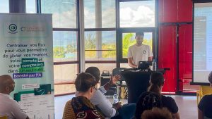 Un homme se tient sur un podium et fait une présentation à un public assis dans une salle bien éclairée, avec un écran et des bannières promotionnelles visibles à l'arrière-plan.