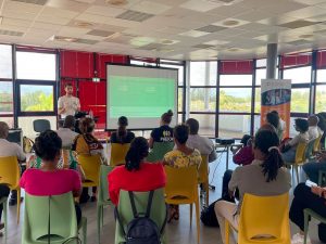 Un groupe de personnes assises sur des chaises colorées écoute un présentateur faire une présentation avec des diapositives dans une salle lumineuse avec de grandes fenêtres.